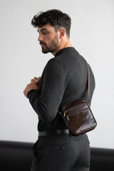 Stylish man in charcoal knitwear and trousers holds a dark brown vertical leather messenger bag against a white studio backdrop
