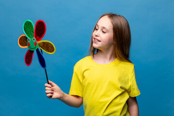 Photo of little cheerful girl in yellow t-shirt, happy positive smile child holds colorful paper toy windmill isolated over blue background. Summer vibes concept. Copy space. High quality photo