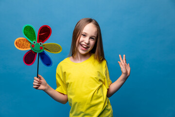 Photo of little cheerful girl in yellow t-shirt, happy positive smile child holds colorful paper toy windmill isolated over blue background. Summer vibes concept. Copy space. High quality photo