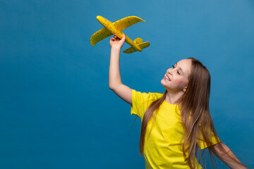 Side view little girl in yellow t-shirt holds a toy airplane on a blue background. Children games and imagination. Copy space. Travel, vacation and dreams of flight concept. High quality photo