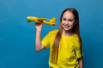 Happy little girl in yellow t-shirt holds a toy airplane on a blue background. Children games and imagination. Copy space. Travel, vacation and dreams of flight concept. High quality photo