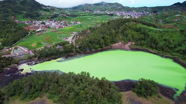 Aerial drone footage of a sulfuric opal color lake in Dieng Plateau, a high altitude volcanic mountain area in Indonesia, with a forest around and villages nearby gardens