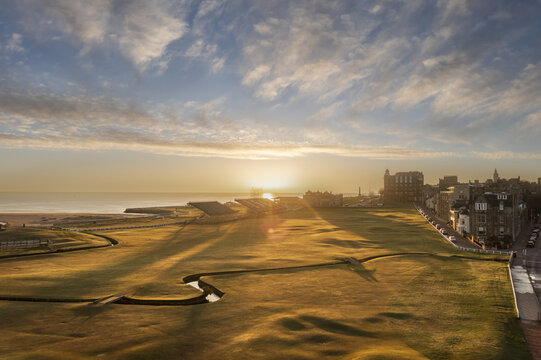 Aerial view of the golden sun casting long shadows over the undulating green landscape of the Old Course, where the Swilcan Burn meanders towards the distant sea, St Andrews, Scotland, United Kingdom.