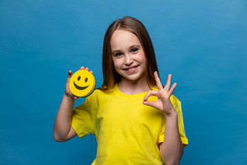 A funny schoolgirl is holding a happy emoji emotion icon in her hands and showing okey sign on blue background. International World smile Day. World emoji day. School Birthday Party. Emotions.