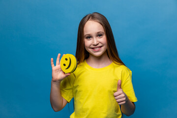 A funny schoolgirl is holding a happy emoji emotion icon in her hands and showing thumbs up sign on blue background. International World smile Day. World emoji day. School Birthday Party. Emotions.