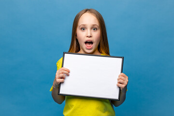 White banner. Surprised shocked Little child schoolgirl in yellow t-shirt on blue background holding white blank paper. Young girl show blank board. Copy space. Mock up. High quality photo