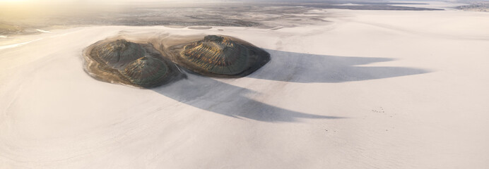Aerial view of twin mounds cast long shadows across a vast, pale expanse, their dark forms contrasting sharply with the surrounding bleached landscape, Karynzharyk, Mangystau Region, Kazakhstan.