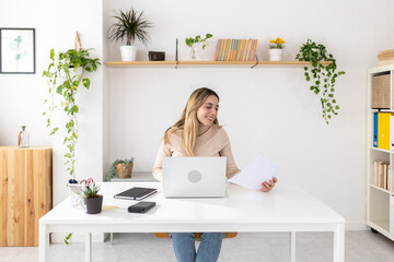 Young adult woman working remotely and reading papers. Relaxed freelancer reviewing documents while typing on her laptop. Creativity and digital work concept.