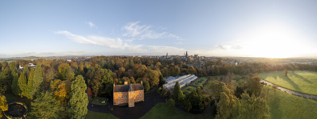 Aerial view of a golden-hued building and glasshouse nestled amidst vibrant autumn foliage, with Dunfermline Abbey standing tall in the distance, Dunfermline, Scotland, United Kingdom.