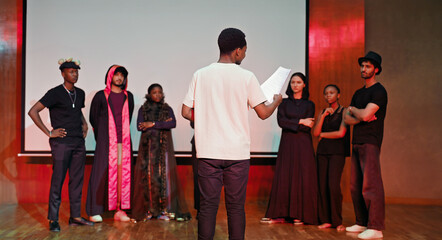 African young adult gen z male man hold scripts paper read play story lines scene standing indoor stage hall. Happy group boy girl clap hand enjoy do work show drama practice day inside campus place