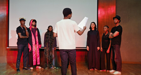 African young adult gen z male man hold scripts paper read play story lines scene standing indoor stage hall. Happy group boy girl clap hand enjoy do work show drama practice day campus place
