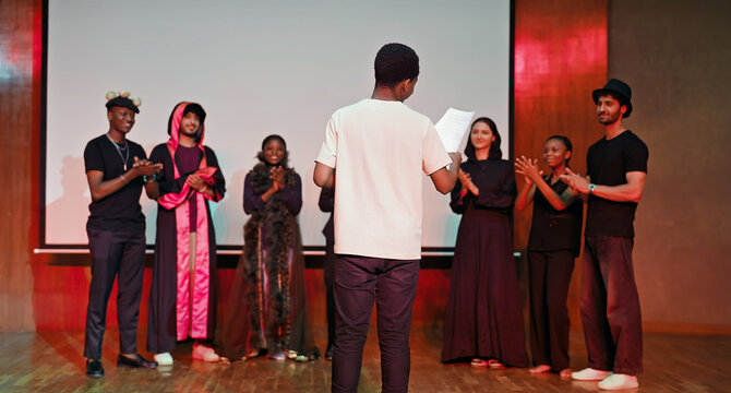 African young adult gen z male man hold scripts paper read play story lines scene standing indoor stage hall. Happy group boy girl clap hand enjoy do work show drama practice day inside campus place
