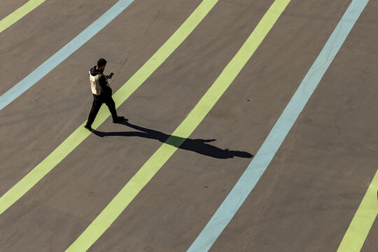View of a lone figure casting a long shadow as he walks across a striped ground, bathed in harsh sunlight, contrasting with the cool blues and greens, Beijing, Beijing, China.