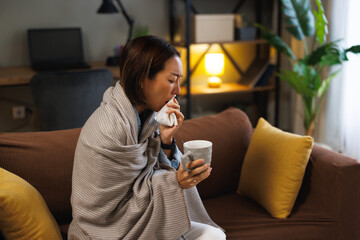 Sick Asian woman blowing nose with tissue and holding mug on couch