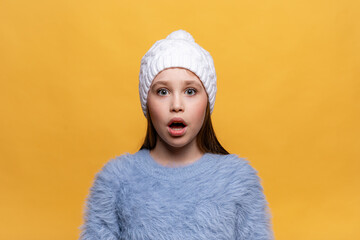 Portrait of little scared girl with eyes wide open, wearing blue sweatshirt, white knitted hat, looking at camera on a yellow background High quality photo. Copy space