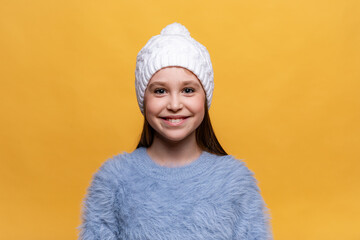 Close up studio portrait of cheerful laughing little girl in blue sweatshirt, white knitted hat on yellow background. Copy space. High quality photo