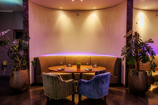 Curved camel banquette with oval table, velvet chairs, and monstera. Title in description. Subtle purple LED wash and warm spots create a cozy lounge mood.