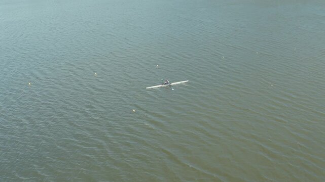 Aerial view of a lone rower cutting through the calm, rippled surface of a vast lake, contrasted against the distant cityscape, Ciudad de la Costa, Departamento de Canelones, Uruguay.