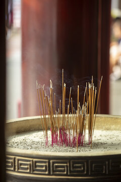 Singapore, Singapore - 12 May 2024: View of incense sticks burning in a decorative pot, smoke curling upwards against the backdrop of a red pillar.