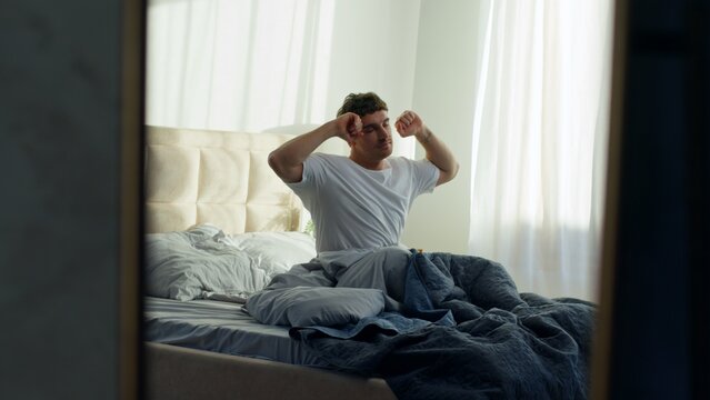 Caucasian man lying bed waking up stretching good morning in bedroom at home sleepy relaxed male guy rubbing eyes after rest comfortable wellbeing awakening new day weekend calm apartment hotel indoor