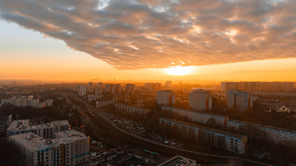 Aerial drone panorama of residential cityscape at golden hour — dramatic cloud shelf over avenue with traffic, apartment blocks and skyline in warm sunset light, European urban landscape, no people