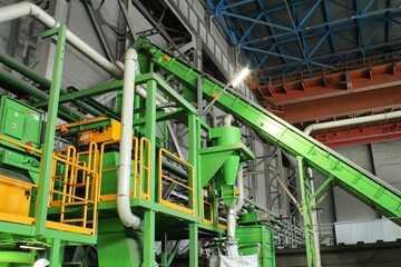 Industrial green conveyor system and recycling machinery in a large factory warehouse with pipes, metal walkways, and heavy duty sorting equipment for waste processing