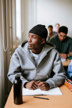 Thoughtful male student wearing do rag looking away while sitting at desk in classroom