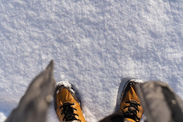 A top-down, personal perspective of feet in warm brown winter boots standing in fresh newly-fallen snow. Top view photo of virgin snow on the ground with person standing in the sunny winter morning.