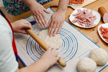 Close-up photo of mother and son cooking pizza together in kitchen. Little boy helping his mother with dinner for family. Mom and child rolling out dough with rolling pin for homemade pepperoni pizza.