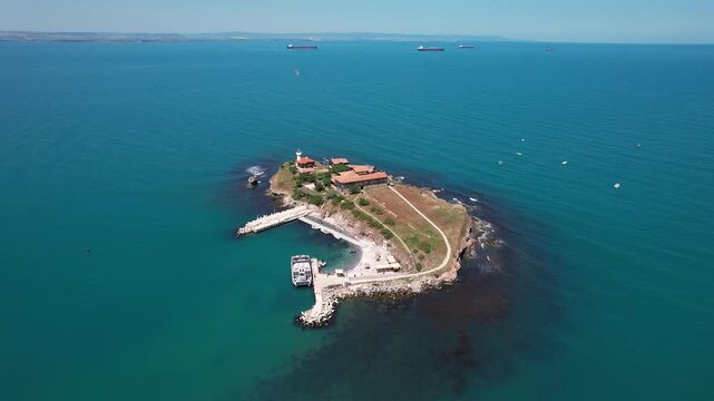 Aerial view of the island with buildings, a lighthouse, and a pier amidst the blue sea with ships sailing in the distance, St. Anastasia Island, Burgas, Bulgaria.