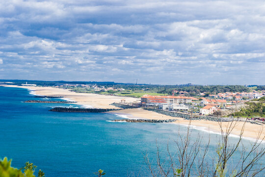 View of the turquoise water meets the sandy beach and buildings under a textured sky, creating a scenic coastal contrast, Anglet, France.