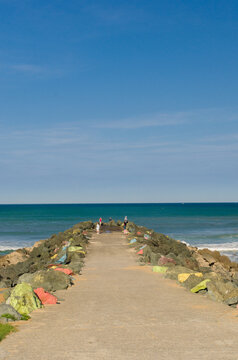 View of a stone pier stretching out into the turquoise sea under a bright blue sky, with people strolling along its length, Anglet, France.