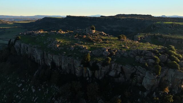 Aerial view of the rugged, rocky plateau with a sheer cliff face contrasting with the softer greens and distant hills, Ittiri, Sardinia, Italy.