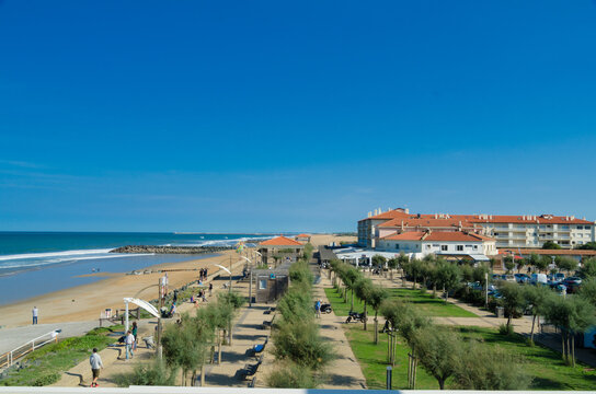 View of golden sands meet azure waters, where people stroll along the beachside path framed by vibrant greenery and a terracotta-roofed building, Anglet, France.