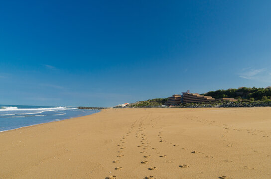 View of golden sand meeting the turquoise ocean under a vast blue sky, leading to a distant architectural structure, Anglet, France.