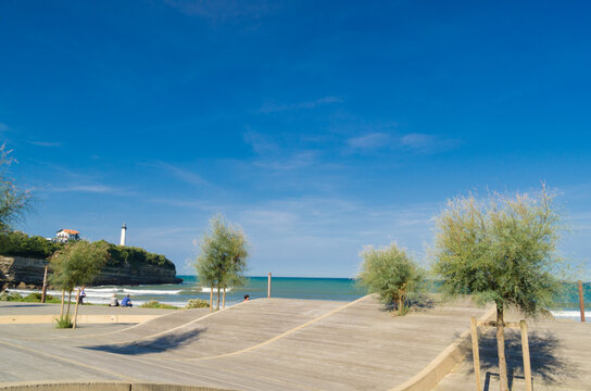 View of bright sunlight illuminating the sandy beach and clear blue ocean with a lighthouse standing tall on the cliff, Anglet, France.