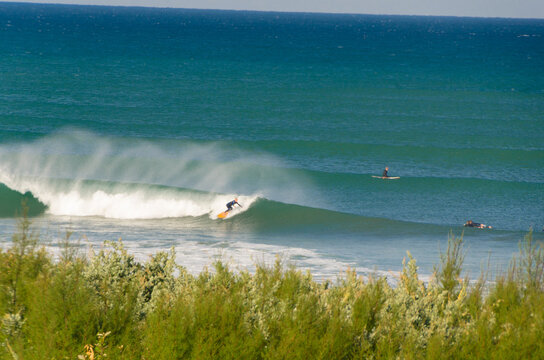 View of a surfer gliding across a turquoise wave under a clear sky as another person paddleboards in the distance, Anglet, France.