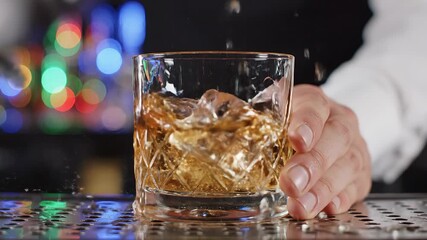 A closeup shot captures a bartenders hand meticulously adding crystalclear ice cubes into a stylish cutglass tumbler filled with rich amber whiskey creating a refreshing splash on a vibrant bar count.