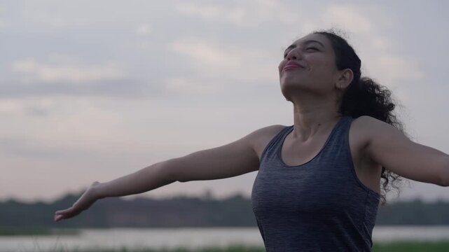 A close-up view of a South Asian woman extending her arms gracefully during a calm lakeside sunrise yoga session