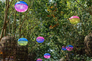 Vibrant Upcycled Lanterns Canopy. Colorful Plastic Food Covers Transformed into Festive Hanging Lamps Amidst Lush Green Tree and Umbrellas. View of Creative Light Fixtures Made From Plastic Food cover