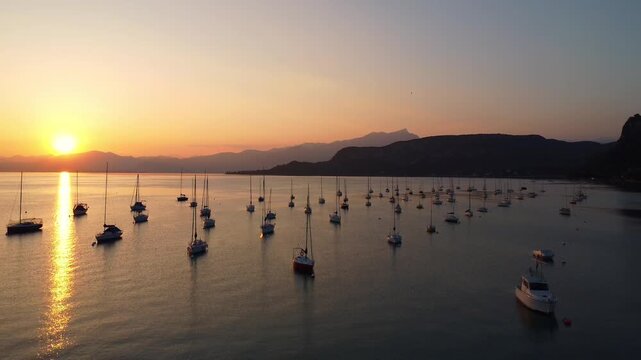 drone view of white sailing yachts anchored in perfect geometric rows on Lake Garda, Italy, under a vivid sunset, reflecting golden light on calm water, evoking elegance and serenity.