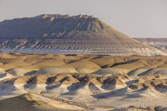 View of sun-drenched, layered mesas rise above the undulating terrain, casting long shadows across the textured ground, Senek, Mangystau Region, Kazakhstan.