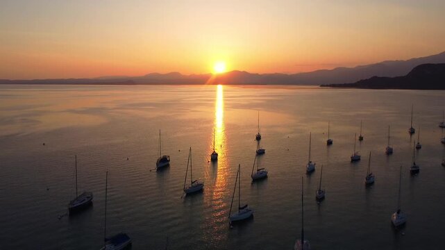 drone view of white sailing yachts anchored in perfect geometric rows on Lake Garda, Italy, under a vivid sunset, reflecting golden light on calm water, evoking elegance and serenity.