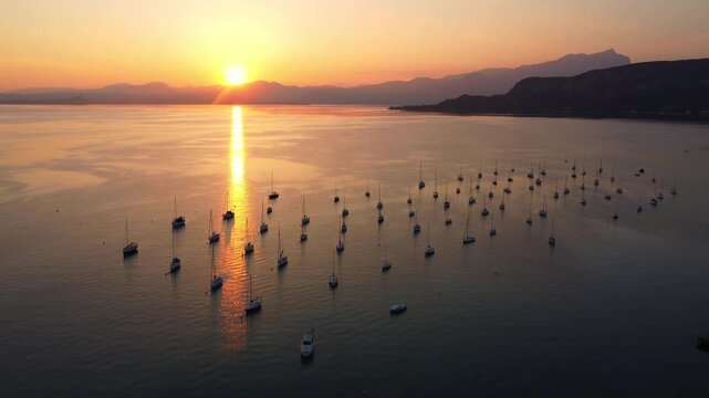 Top-down drone video of white sailing yachts anchored in perfect geometric rows on Lake Garda, Italy, under a vibrant sunset, showcasing symmetry, calm water, and luxury lakeside atmosphere