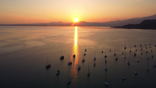 Top-down drone video of white sailing yachts anchored in perfect geometric rows on Lake Garda, Italy, under a vibrant sunset, showcasing symmetry, calm water, and luxury lakeside atmosphere