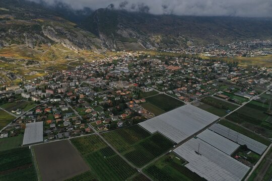 Aerial view of a sprawling town nestled beside towering mountains, with agricultural fields creating a patchwork of greens and whites under a moody sky, Zermatt, Valais, Switzerland.
