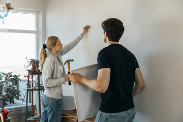 Young man holding picture frame while looking at girlfriend hammering nail on wall