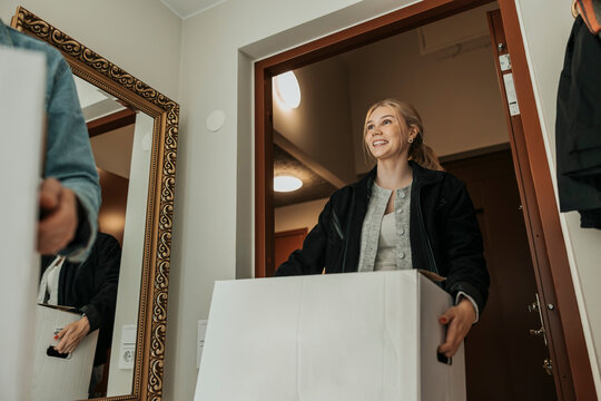 Low angle view of smiling young woman carrying cardboard box walking through doorway of new apartment