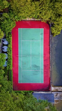 Aerial view of a vibrant tennis court surrounded by lush greenery and dark water, a striking contrast of red and green, Hunters Hill, New South Wales, Australia.