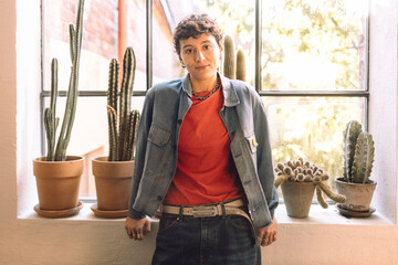 Portrait of confident short haired woman standing near cactus plants kept on window sill